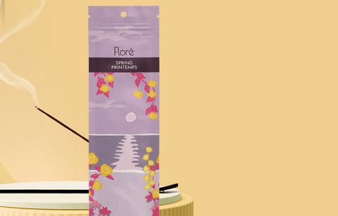 A pale purple package of incense sticks that reads Flore Spring with an image of yellow flowers framing a purple lake with a grey sun in the sky. The package stands upright on a podium next to a burning incense stick against a pale golden background.