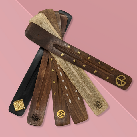 Six long flat wooden incense burners of varying shades from dark wood to light, arranged in a fan configuration. Each incense burner has a different design or decal, including a square gold stamp, lotus flower, ohm symbol, diamond pattern, tree of life and a peace symbol.