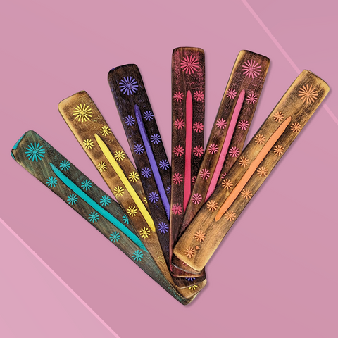 Six long flat wooden incense burners each featuring a different colour floral design laying side by side diagonally on a pink background. From left to right the colours of the incense burners are teal, yellow, blue, dark pink, light pink, peach.