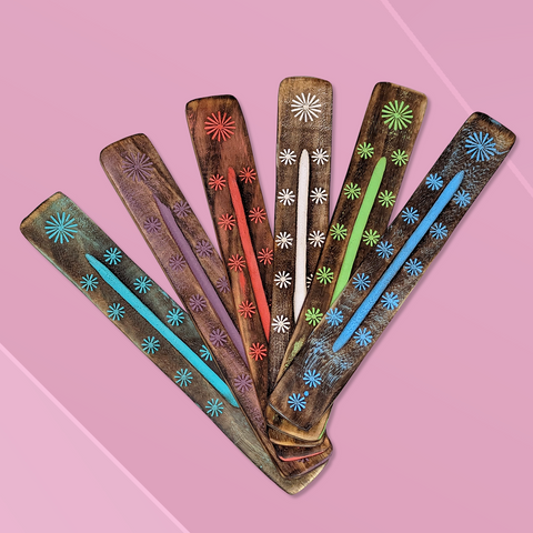 Six long flat wooden incense burners each featuring a different colour floral design laying side by side diagonally on a pink background. From left to right the colours of the incense burners are baby blue, mauve, red, white, sage green, blue.