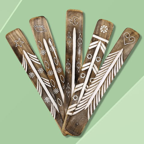 Five long flat wooden incense burners each with unique white painted carvings arranged in a fan formation against a mossy green background.