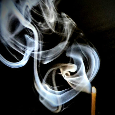 Spirals of white incense smoke coming from the tip of an incense stick burning against a black background.