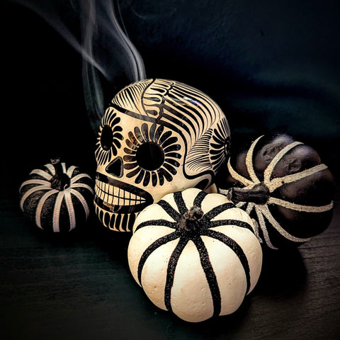 A black and white sugar skull incense burner with smoke coming out of its eye sockets, surrounded by black and white striped pumpkins.