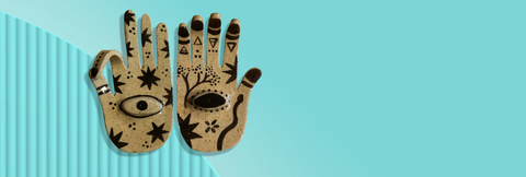 An overhead view of two ceramic incense burners shaped like hands against a pale turquoise background. The right hand is facing palm up and each finger has one of the triangle element symbols for earth, air, fire and water, and the palm is painted with floral images, a snake, and a third eye in the centre of the palm. The left hand is also facing palm up with the index finger and thumb touching, and is painted with stars and a third eye in the centre of the palm. Original design by Toronto illustrator Emily