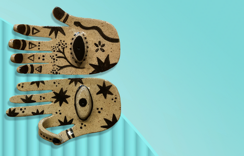 An overhead view of two ceramic incense burners shaped like hands against a pale turquoise background. The right hand is facing palm up and each finger has one of the triangle element symbols for earth, air, fire and water, and the palm is painted with floral images, a snake, and a third eye in the centre of the palm. The left hand is also facing palm up with the index finger and thumb touching, and is painted with stars and a third eye in the centre of the palm. Original design by Toronto illustrator Emily