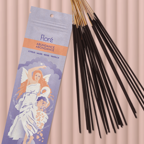 A pale purple package of incense sticks that reads Flore Abundance with an image of an angelic woman holding a cornucopia. The package lies with a bundle of incense sticks on a rosy beige background.