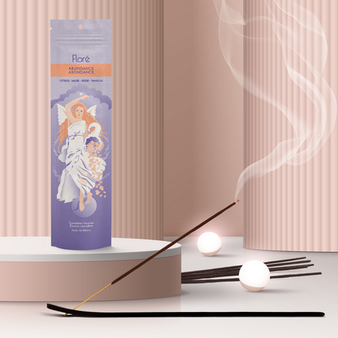 A pale purple package of incense sticks that reads Flore Abundance with an image of an angelic woman holding a cornucopia. The package stands upright on a podium next to a burning incense stick and white orbs against a rosy beige background.