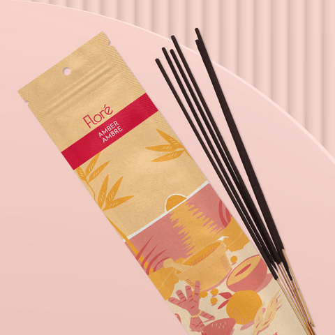 A pale orange package of incense sticks that reads Flore Amber with an image of red and orange fruits, spices and mortar and pestle on a beach at sunset. The package lies with a bundle of incense sticks on a pale peach background.