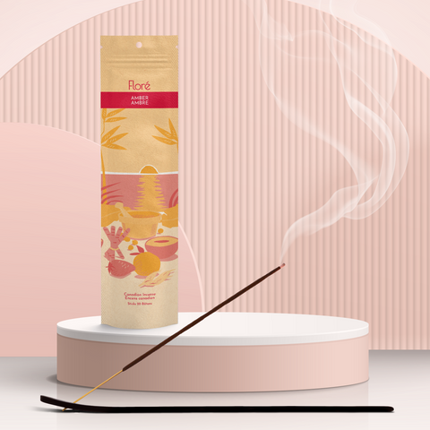 A pale orange package of incense sticks that reads Flore Amber with an image of red and orange fruits, spices and mortar and pestle on a beach at sunset. The package stands upright on a podium next to a burning incense stick against a pale peach background.