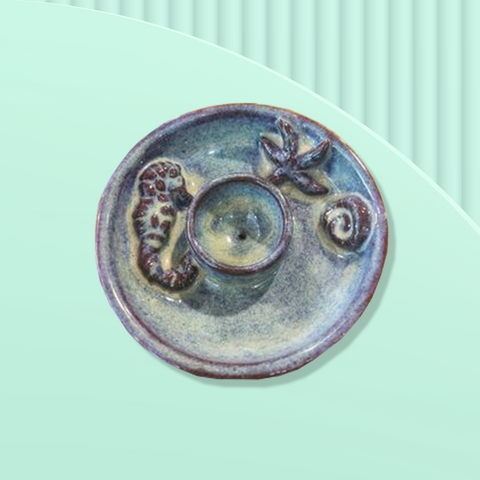 Overhead view of a faded blue round ceramic incense burner dish with a relief sculpture seahorse, a star fish and seashell. The incense burner sitting on a pale seafoam green background. 