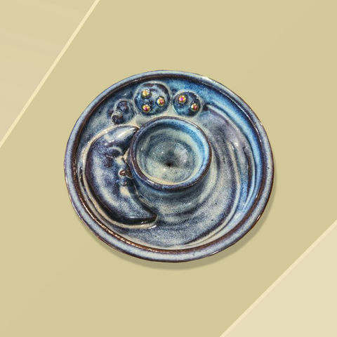 Overhead view of a dark blue round ceramic incense burner dish with a relief sculpture of a crescent moon and several planets with silver balls. The incense burner is sitting on a sandy coloured background.