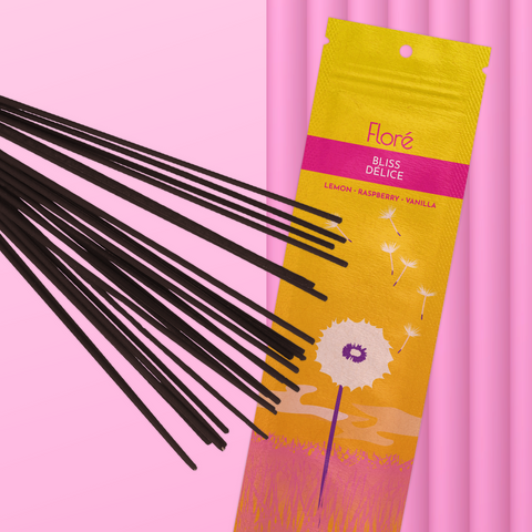 A golden orange package of incense sticks that reads Flore Bliss with an image of a single white dandelion in a pink grass meadow with fluffy white seeds drifting into a golden sky.The package lies with a bundle of incense sticks on a pink background.