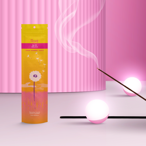 A golden orange package of incense sticks that reads Flore Bliss with an image of a single white dandelion in a pink grass meadow with fluffy white seeds drifting into a golden sky. The package stands upright next to a burning incense stick and white orbs against a pink background.