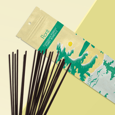 A pale yellow package of incense sticks that reads Flore Canadian Forest with an image of pine trees growing on the shore of a golden lake with a yellow sun in the sky. The package lies with a bundle of incense sticks on a pale yellow-green background.