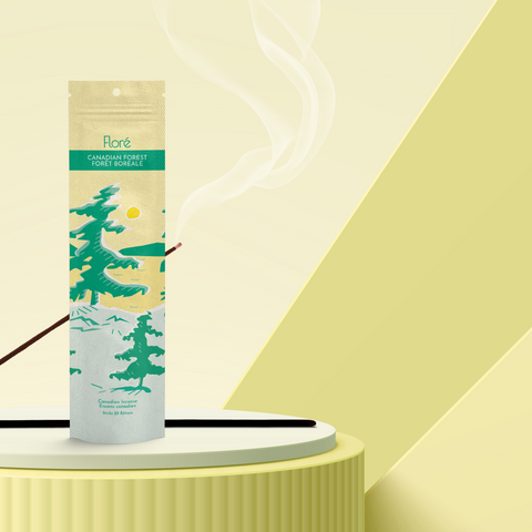 A pale yellow package of incense sticks that reads Flore Canadian Forest with an image of pine trees growing on the shore of a golden lake with a yellow sun in the sky. The package stands upright on a podium next to a burning incense stick against a pale yellow-green background.