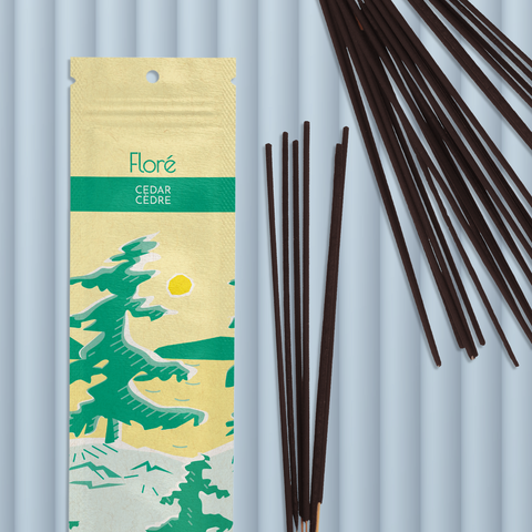 A pale yellow package of incense sticks that reads Flore Cedar with an image of pine trees growing on the shore of a golden lake with a yellow sun in the sky. The package lies with a bundle of incense sticks on a steely grey background.