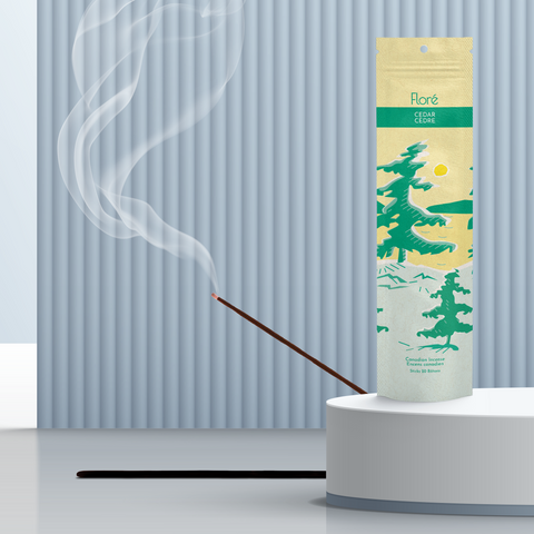 A pale yellow package of incense sticks that reads Flore Cedar with an image of pine trees growing on the shore of a golden lake with a yellow sun in the sky. The package stands upright on a podium next to a burning incense stick against a steely grey background.