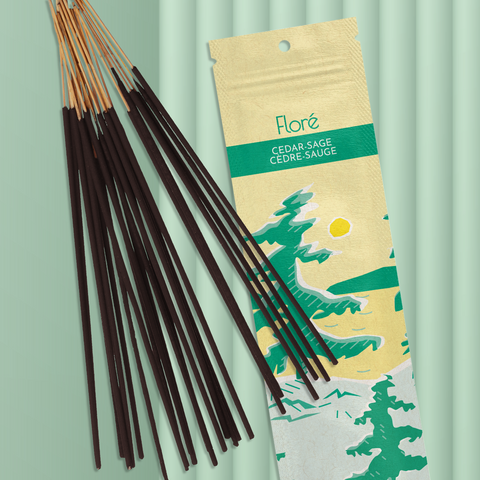 A pale yellow package of incense sticks that reads Flore Cedar-Sage with an image of pine trees growing on the shore of a golden lake with a yellow sun in the sky. The package lies with a bundle of incense sticks on a pale green background.