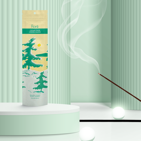 A pale yellow package of incense sticks that reads Flore Cedar-Sage with an image of pine trees growing on the shore of a golden lake with a yellow sun in the sky. The package stands upright on a podium next to a burning incense stick and white orbs against a pale green background.