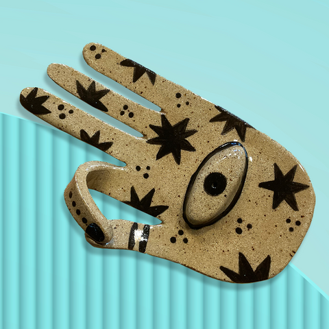 An overhead view of a ceramic incense burner shaped like a left hand against a pale turquoise background. The hand is facing palm up with the index finger and thumb touching, and is painted with stars and a third eye in the centre of the palm. Original design by Toronto illustrator Emily Elise.