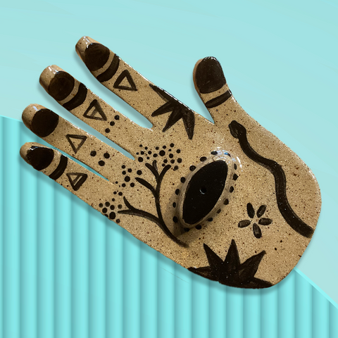 An overhead view of a ceramic incense burner shaped like a right hand against a pale turquoise background. The hand is facing palm up and each finger has one of the triangle element symbols for earth, air, fire and water, and the palm is painted with floral images, a snake, and a third eye in the centre of the palm. Original design by Toronto illustrator Emily Elise.