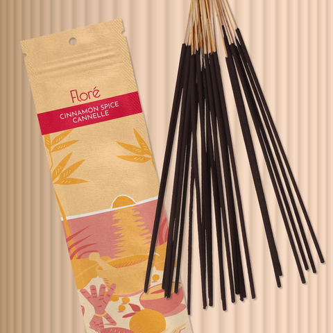 A pale orange package of incense sticks that reads Flore Cinnamon Spice with an image of red and orange fruits, spices and mortar and pestle on a beach at sunset. The package lies with a bundle of incense sticks on a taupe background.