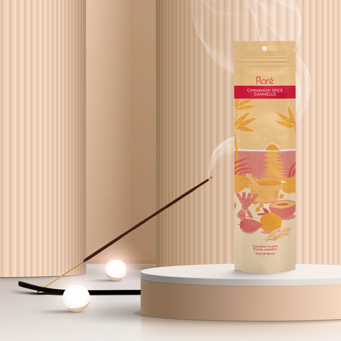 A pale orange package of incense sticks that reads Flore Cinnamon Spice with an image of red and orange fruits, spices and mortar and pestle on a beach at sunset. The package stands upright on a podium next to a burning incense stick and white orbs against a taupe background.