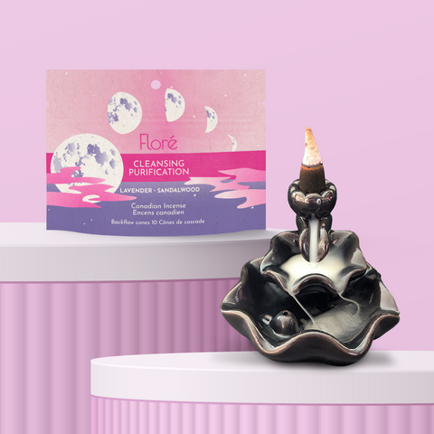 A package of backflow incense cones that reads Flore Cleansing with images of moon phases against a misty pink and purple background. The package stands upright on a podium next to a burning incense cone on a backflow cone burner against a pale pink background. The incense smoke is spilling down the burner creating a waterfall of smoke. 