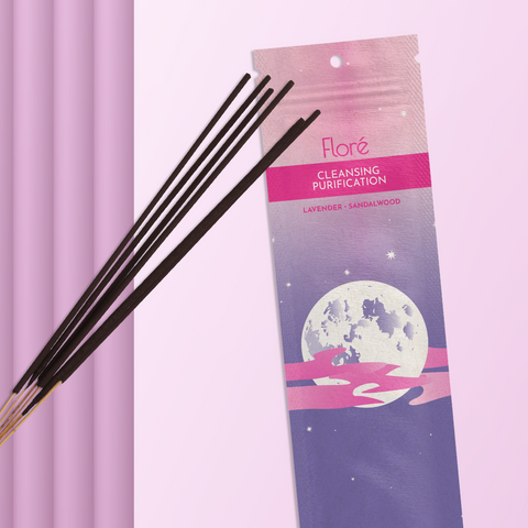 A purple package of incense sticks that reads Flore Cleansing with an image of a full moon surrounded by a jagged pink cloud in a starry purple night sky. The package lies with a bundle of incense sticks on a pale pinky purple background.