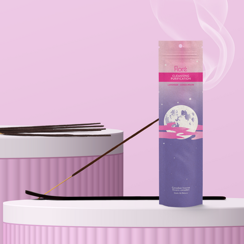 A purple package of incense sticks that reads Flore Cleansing with an image of a full moon surrounded by a jagged pink cloud in a starry purple night sky. The package stands upright on a podium next to a burning incense stick against a pale pinky purple background.