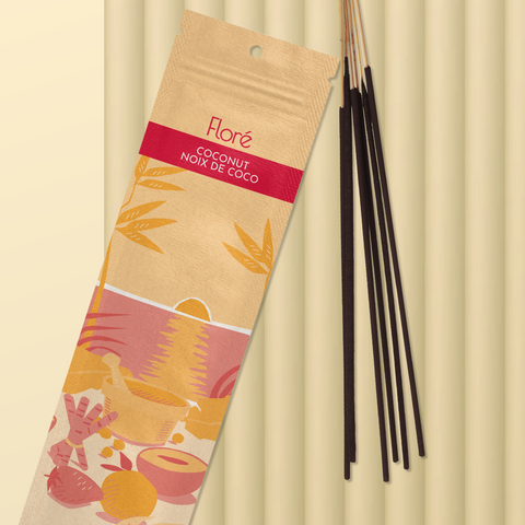 A pale orange package of incense sticks that reads Flore Coconut with an image of red and orange fruits, spices and mortar and pestle on a beach at sunset. The package lies with a bundle of incense sticks on a pale yellow background.