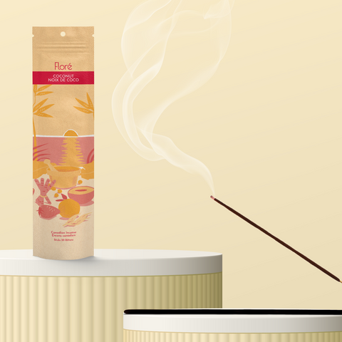 A pale orange package of incense sticks that reads Flore Coconut with an image of red and orange fruits, spices and mortar and pestle on a beach at sunset. The package stands upright on a podium next to a burning incense stick against a pale yellow background.