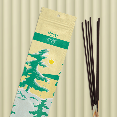 A pale yellow package of incense sticks that reads Flore Cypress with an image of pine trees growing on the shore of a golden lake with a yellow sun in the sky. The package lies with a bundle of incense sticks on a pale greeny taupe background.