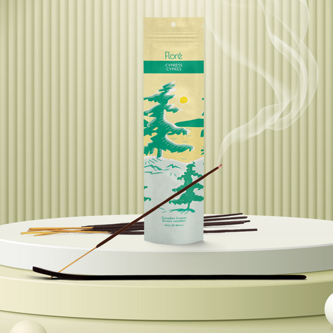 A pale yellow package of incense sticks that reads Flore Cypress with an image of pine trees growing on the shore of a golden lake with a yellow sun in the sky. The package stands upright on a podium next to a burning incense stick against a pale greeny taupe background.