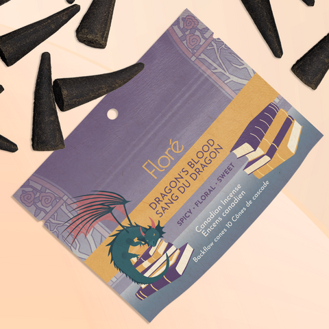 A package of backflow incense cones that reads Flore Dragon's Blood with an image of a dragon, piles of books and stained glass windows against a grey purple background. The package lies with a bunch of incense cones on a pale gold background.