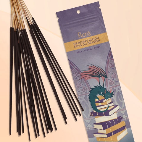 A smoky purple package of incense sticks that reads Flore Dragon’s Blood with an image of a dragon resting on top of a pile of books in front of gothic floral stained glass windows. The package lies with a bundle of incense sticks on a pale golden peach background.