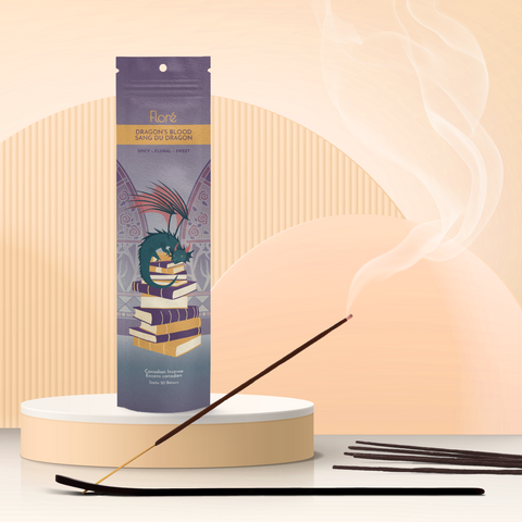 A smoky purple package of incense sticks that reads Flore Dragon’s Blood with an image of a dragon resting on top of a pile of books in front of gothic floral stained glass windows. The package stands upright on a podium next to a burning incense stick against a pale golden peach background.