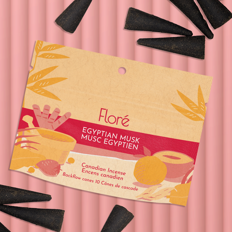 A pale orange package of backflow incense cones that reads Flore Egyptian Musk with an image of red and orange fruits, spices and mortar and pestle on a beach at sunset. The package lies with a bunch of incense cones on a pale red background.