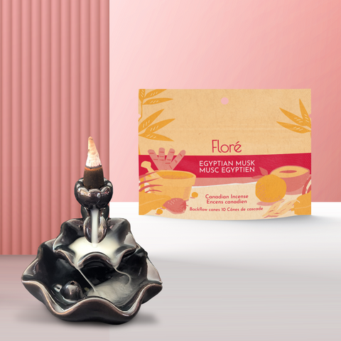 A pale orange package of backflow incense cones that reads Flore Egyptian Musk with an image of red and orange fruits, spices and mortar and pestle on a beach at sunset. The package stands upright next to a burning incense cone on a backflow cone burner against a pale red background. The incense smoke is spilling down the burner creating a waterfall of smoke. 