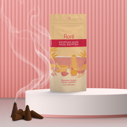 A pale orange package of incense cones that reads Flore Egyptian Musk with an image of red and orange fruits, spices and mortar and pestle on a beach at sunset. The package stands upright next to a burning incense cone against a pale red background.
