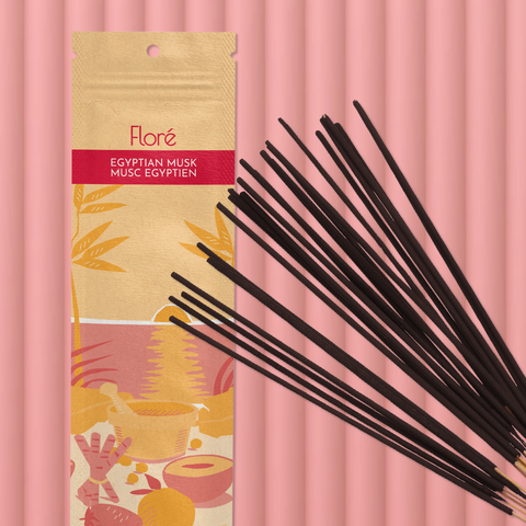 A pale orange package of incense sticks that reads Flore Egyptian Musk with an image of red and orange fruits, spices and mortar and pestle on a beach at sunset. The package lies with a bundle of incense sticks on a pale red background.