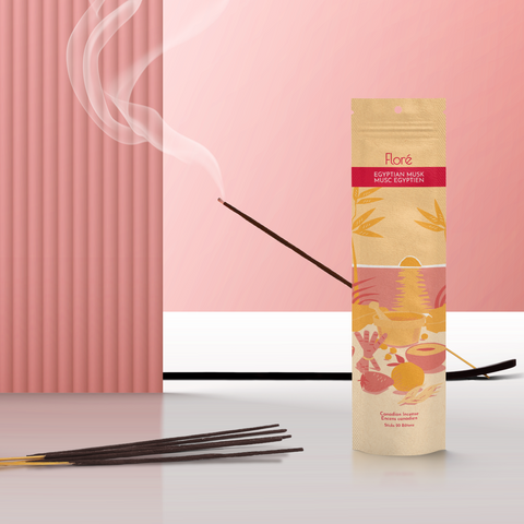 A pale orange package of incense sticks that reads Flore Egyptian Musk with an image of red and orange fruits, spices and mortar and pestle on a beach at sunset. The package stands upright next to a burning incense stick against a pale red background.