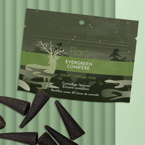 A dark green package of backflow incense cones that reads Flore Evergreen with an image of a deer silhouette with antlers that look like tree branches against a dark forest and starry night sky. The package lies with a bunch of incense cones on a mossy green background.