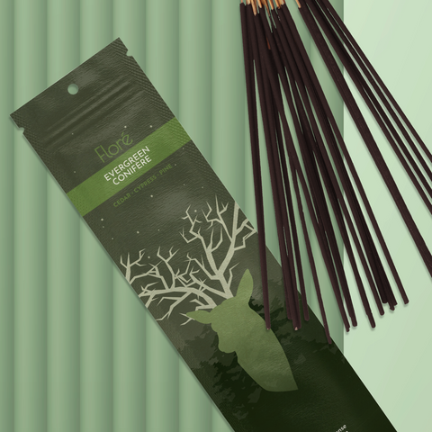 A dark green package of incense sticks that reads Flore Evergreen with an image of a deer head silhouette with antlers that look like tree branches against a dark forest and starry night sky. The package lies with a bundle of incense sticks on a mossy green background.