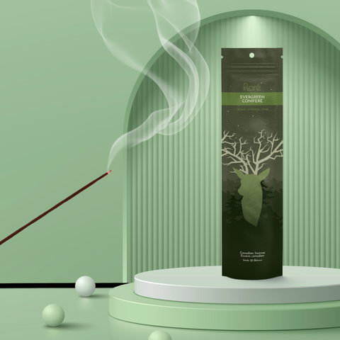 A dark green package of incense sticks that reads Flore Evergreen with an image of a deer head silhouette with antlers that look like tree branches against a dark forest and starry night sky. The package stands upright on a podium next to a burning incense stick against a mossy green background.