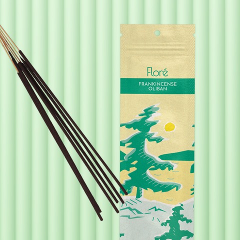 A pale yellow package of incense sticks that reads Flore Frankincense with an image of pine trees growing on the shore of a golden lake with a yellow sun in the sky. The package lies with a bundle of incense sticks on a minty green background.