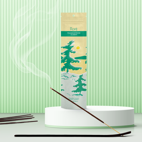 A pale yellow package of incense sticks that reads Flore Frankincense with an image of pine trees growing on the shore of a golden lake with a yellow sun in the sky. The package stands upright on a podium next to a burning incense stick against a minty green background.