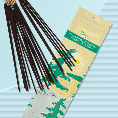 A pale yellow package of incense sticks that reads Flore Frankincense and Myrrh with an image of pine trees growing on the shore of a golden lake with a yellow sun in the sky. The package lies with a bundle of incense sticks on a pale blue background.