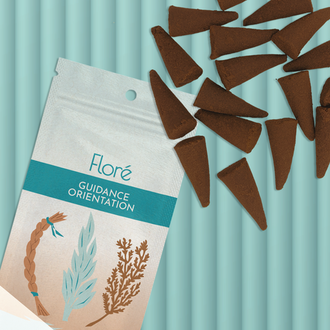 A pale blue-grey package of incense cones that reads Flore Guidance with an image of a sweetgrass braid, sage leaves and a cedar bough. The package lies with a bunch of incense cones on a pale aqua background.