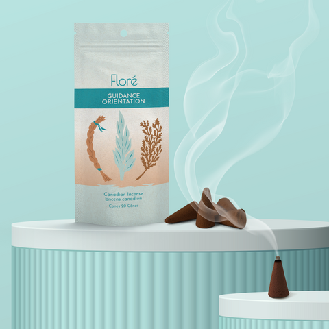 A pale blue-grey package of incense cones that reads Flore Guidance with an image of a sweetgrass braid, sage leaves and a cedar bough. The package stands upright on a podium next to a burning incense cone against a pale aqua background.